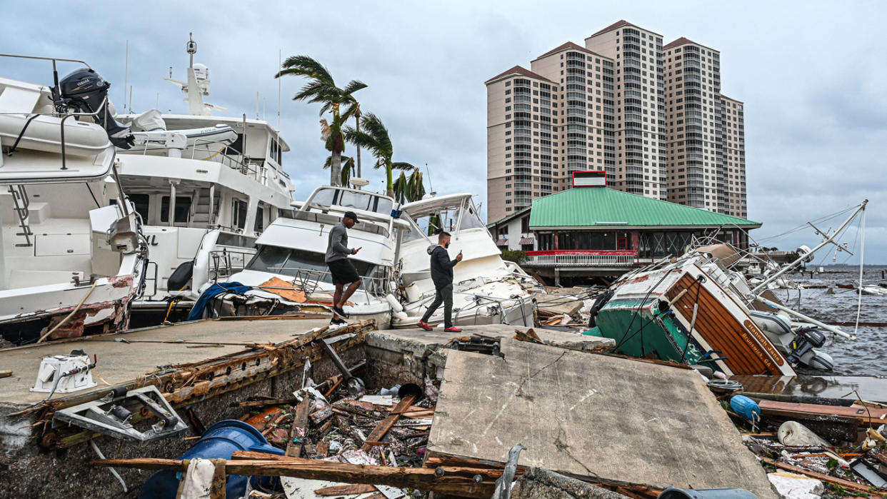 Two men walk along a heavily damaged dock near a few palm trees among half a dozen upended boats and floating debris.