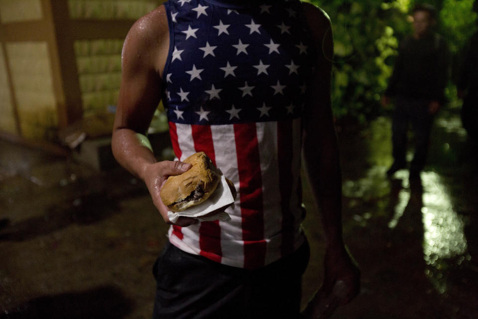 In this Oct. 16, 2018 photo, a Honduran migrant wearing a U.S. flag motif shirt, holds a sandwich at an improvised shelter in Chiquimula, Guatemala. U.S. President Donald Trump threatened on Tuesday to cut aid to Honduras if it doesn't stop the impromptu caravan of migrants, but it remains unclear if governments in the region can summon the political will to physically halt the determined border-crossers. (AP Photo/Moises Castillo)