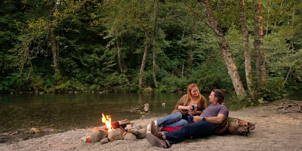 Mel and Jack sitting next to a river as a small logfire burns.