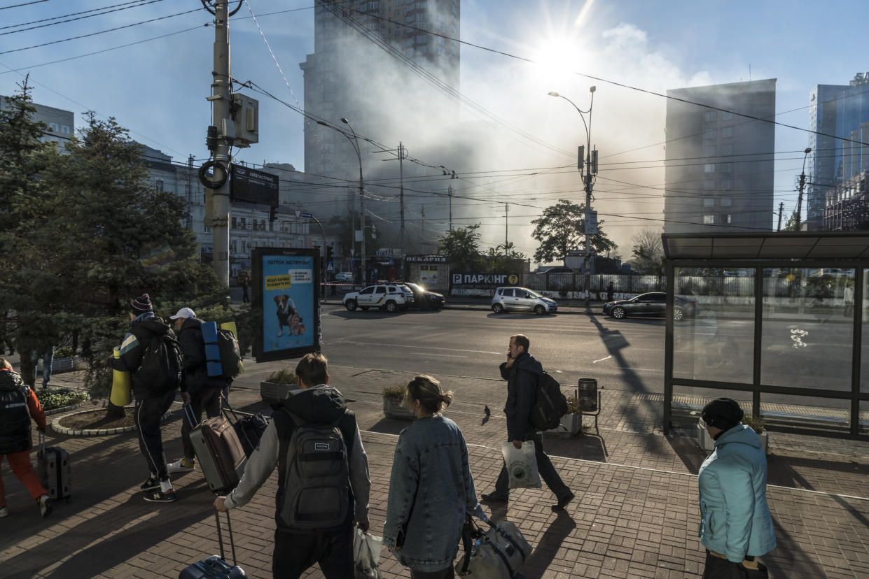 Unas nubes de humo salen del sitio donde sucedió un ataque con drones mientras pasajeros caminan con su equipaje desde unas estaciones de trenes y autobuses cercanas en Kiev, Ucrania, el 17 de octubre de 2022. (Brendan Hoffman/The New York Times)
