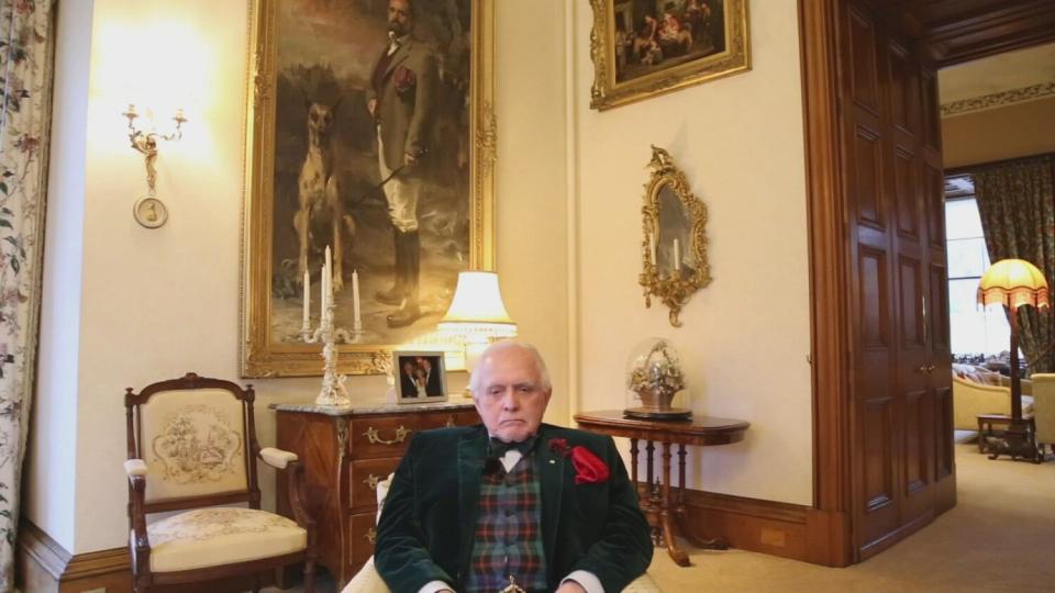 A man sits inside a room in a castle, in front of a large painting.