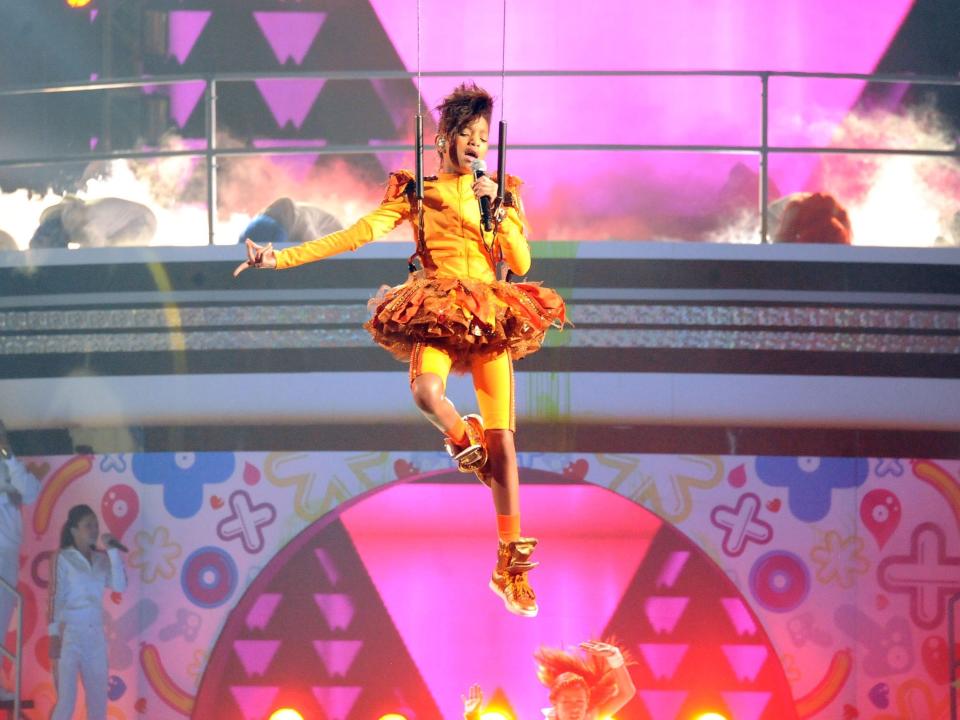 Young Willow Smith is suspended in the air by a harness. She's wearing an all orange get-up (from shoes to track suit to matching tutu). Behind her is a brightly lit, colorful stage.