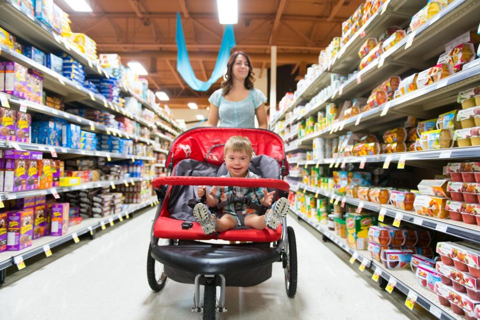 A parent leads a smiling child through a grocery store while scanning the shelves for items to purchase.