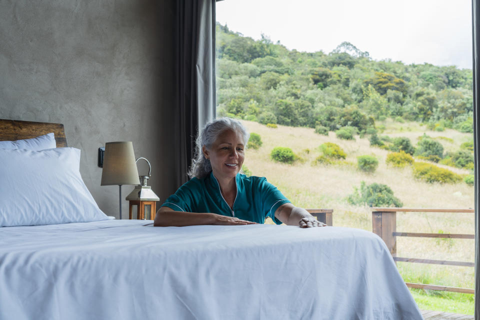 An elderly woman with long gray hair, wearing a teal top, smiling and making a bed in a room with a large window showcasing a scenic, green landscape