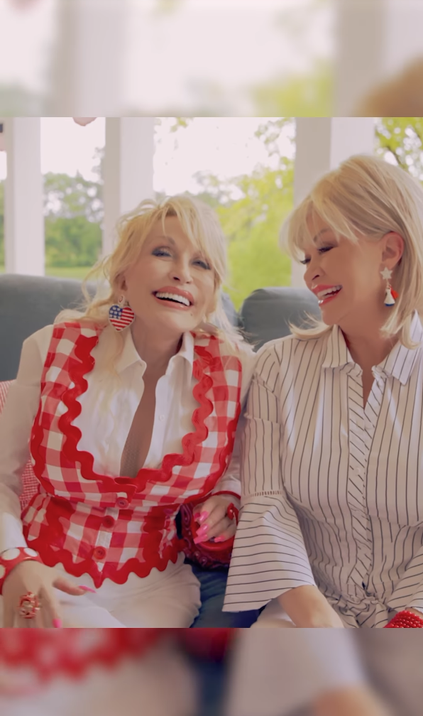 Dolly Parton and another woman sit and laugh. Dolly is wearing a red gingham vest with a white blouse, while the other woman is in a striped blouse