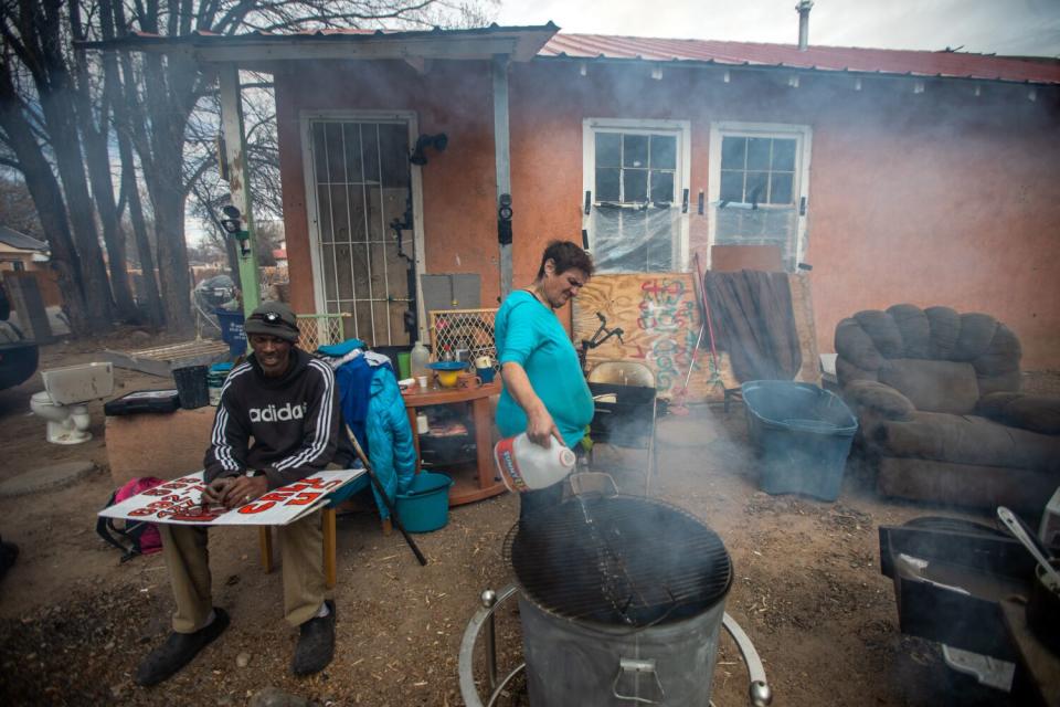 A woman pours fluid into a fire inside a large barrel as a man sits next to her