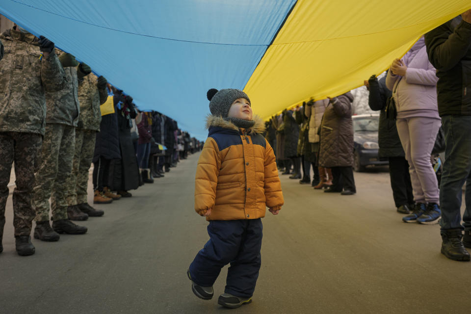 A child walks under a large Ukrainian flag carried by people marking a "day of unity" in Sievierodonetsk, the Luhansk region, eastern Ukraine, Wednesday, Feb. 16, 2022. The flags celebrated survival, endurance and above all, defiance. One blue and yellow banner curved along the edge of a stadium field. Others were tiny handheld things. One made it on the Olympic podium. Ukraine's president declared Wednesday a day of national unity in the face of “hybrid threats.” (AP Photo/Vadim Ghirda)