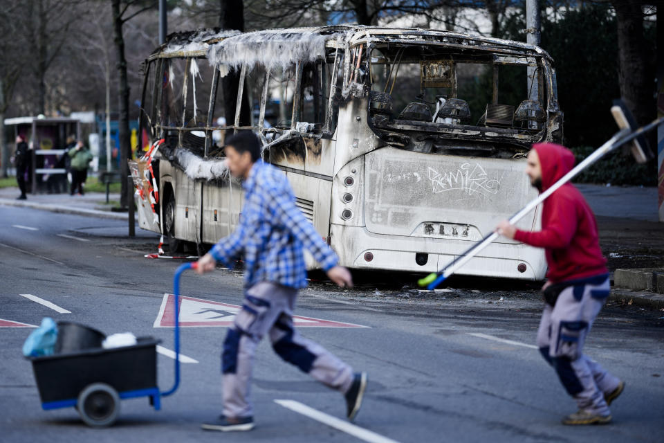 Two men cross the street in front of a burned-out bus standing beneath a residential building, in the district Neukoelln in Berlin, Germany, Tuesday, Jan. 3, 2023. People across Germany on Saturday resumed their tradition of setting off large numbers of fireworks in public places to see in the new year. The bus was set on fire during the New Year's celebrations. (AP Photo/Markus Schreiber)