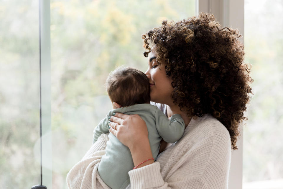 A woman holding a baby close to her chest while gently kissing the baby's head, standing by a window with foliage visible outside