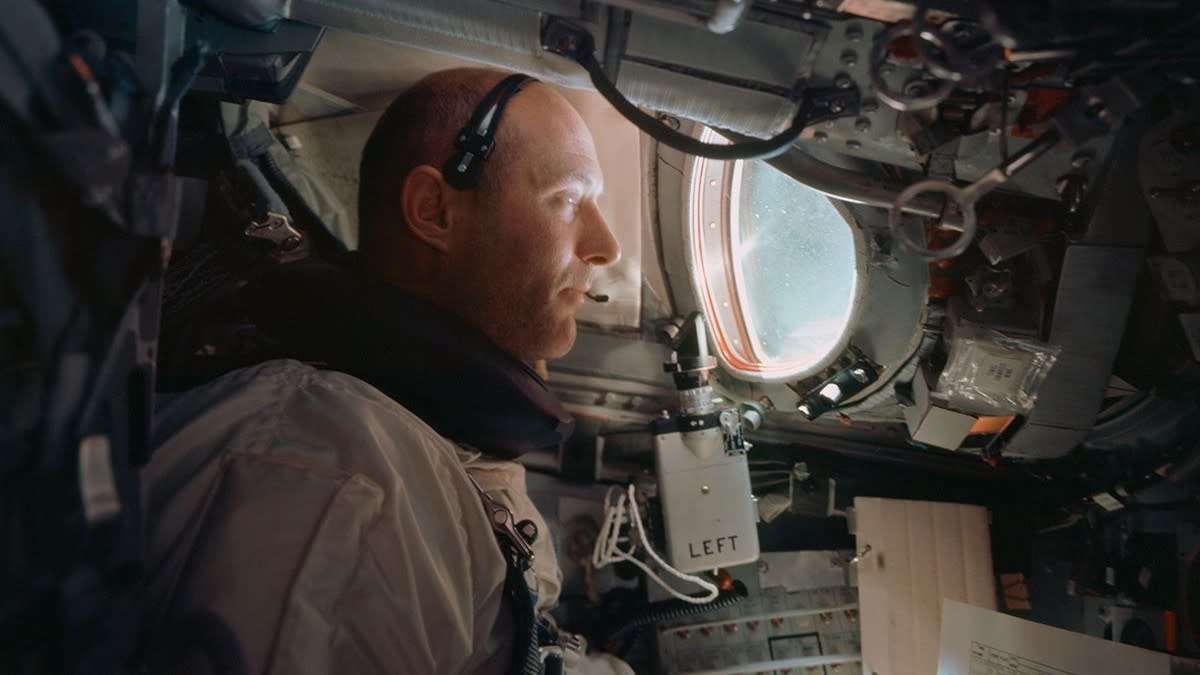  A man in a white flight suit inside a cramped spacecraft capsule filled with screens and wires. 