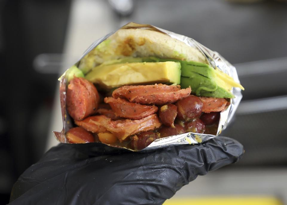 In this June 11, 2019 photo, a food server shows a Venezuelan arepa made with Colombian ingredients at the "Arepas Cafe" fast food restaurant in Bogota, Colombia. Venezuelans like to jest that their beloved arepas are so widely consumed that babies come out of the womb with the corn flatbreads already in hand. Now, as millions flee their homeland's turmoil, they are taking Venezuela's most ubiquitous dish with them.(AP Photo/Fernando Vergara)