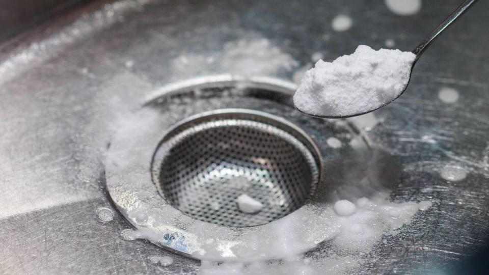 How to unclog kitchen sink: From a high angle view, a woman's hand is adding a mixture of sodium bicarbonate, vinegar, and hot water into a kitchen sink, down the drain.