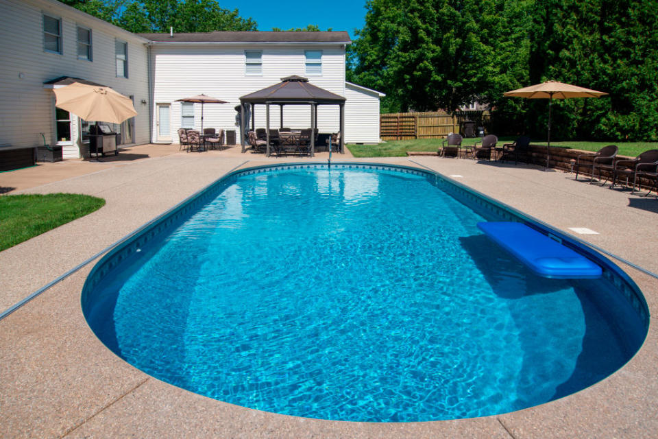 Backyard with an in-ground swimming pool surrounded by lounge chairs, umbrellas and a gazebo. A large white house can be seen in the background