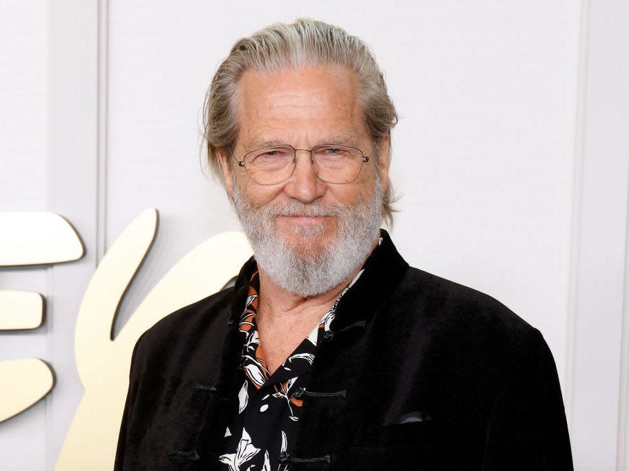 jeff bridges wearing a patterned shirt and black suit jacket, smiling on a red carpet. he's wearing thin rimmed glasses, and his hair is slicked back, his silver beard neatly trimmed