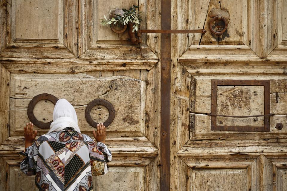 A woman prays in front of the closed Church of the Holy Sepulchre, a place where Christians believe Jesus Christ was buried, as a palm hangs on the door, in Jerusalem's Old City, Sunday, April 5, 2020. The traditional Palm Sunday procession was cancelled due to restrictions imposed to contain the spread of the coronavirus. (AP Photo/Ariel Schalia