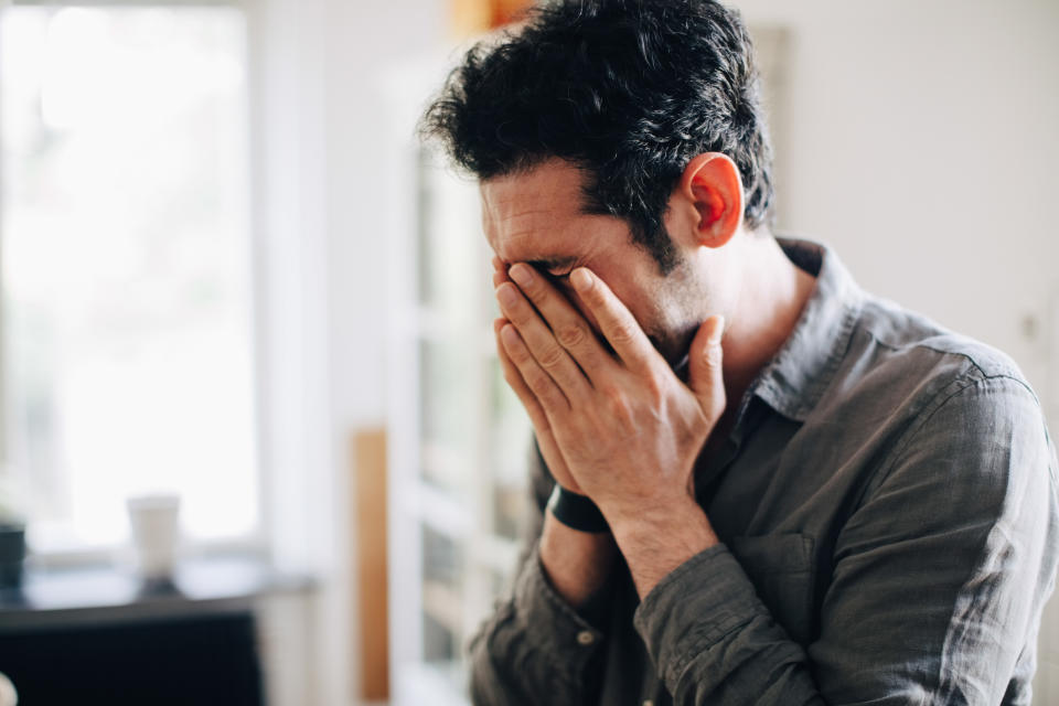 A person in a casual shirt stands indoors, covering their face with both hands in an emotional or stressed gesture