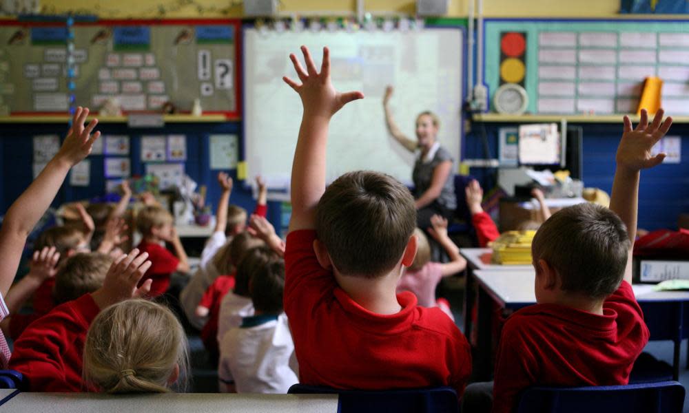 Children in a classroom raising their hands, as teacher also raises hers.