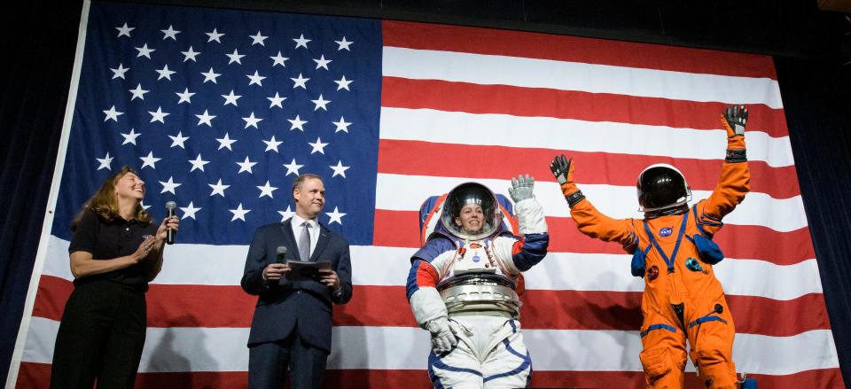 person wearing spacesuit stands with two others in front of USA flag