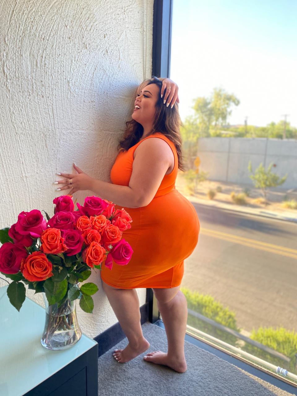 Julia wearing a body-conscious dress poses next to a wall and in front of a vase of roses