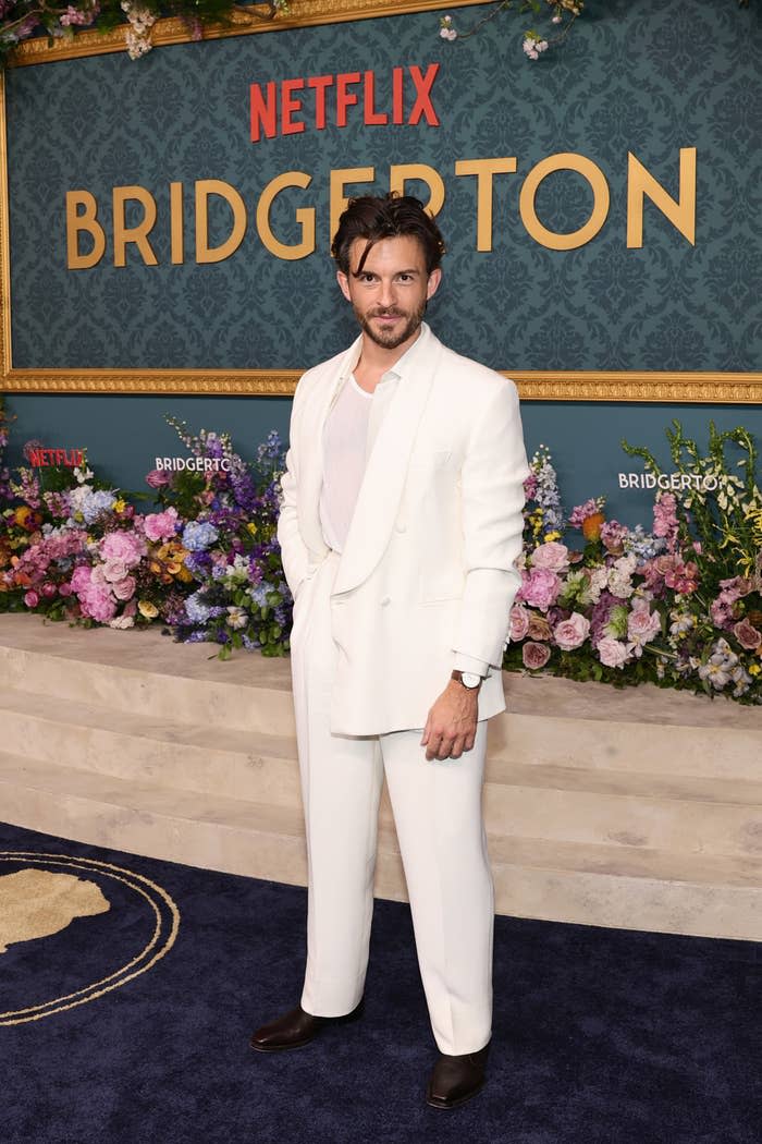 A man in a white suit stands on the Netflix "Bridgerton" the red carpet of the event, decorated with floral arrangements