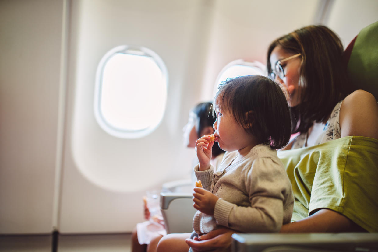 Little toddler girl having snacks joyfully while sitting on her mom’s lap on the airplane