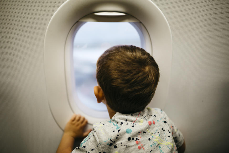 A young child looks out an airplane window, peering at the view outside. The child wears a patterned shirt and holds onto the window's frame