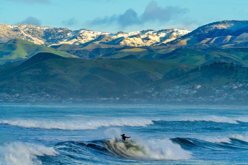 A surfer rides a wave Thursday, Feb. 23, 2023, on the opening morning of the SLO CAL Open competition in Morro Bay, as the sun illuminates the snowy Santa Lucia Range above Cayucos in the distance.