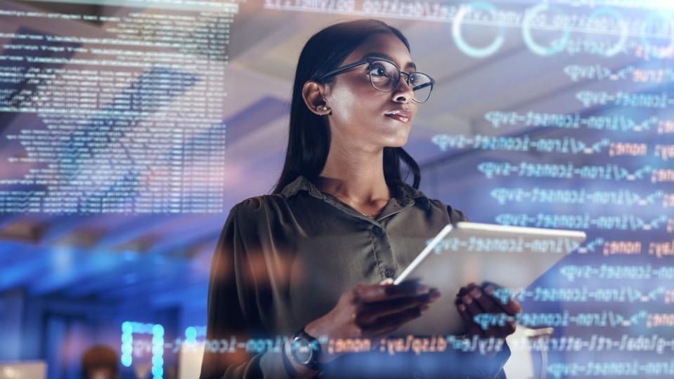 A woman holds a tablet and looks at numbers and symbols on a virtual screen.