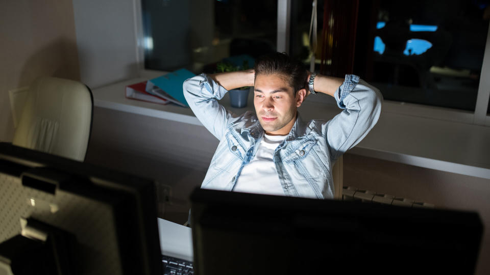 High angle portrait of handsome man chilling while using computer sitting at desk in dark office late at night.