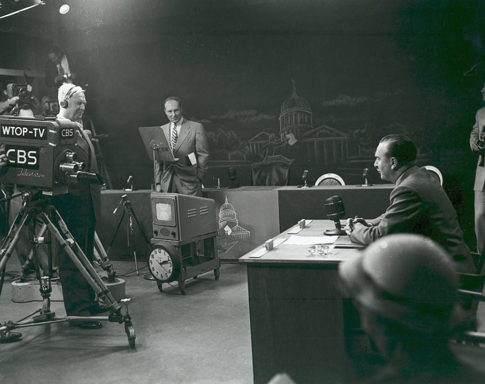 A vintage TV studio scene with a news anchor seated at a desk, a cameraman filming, and another person standing near a podium