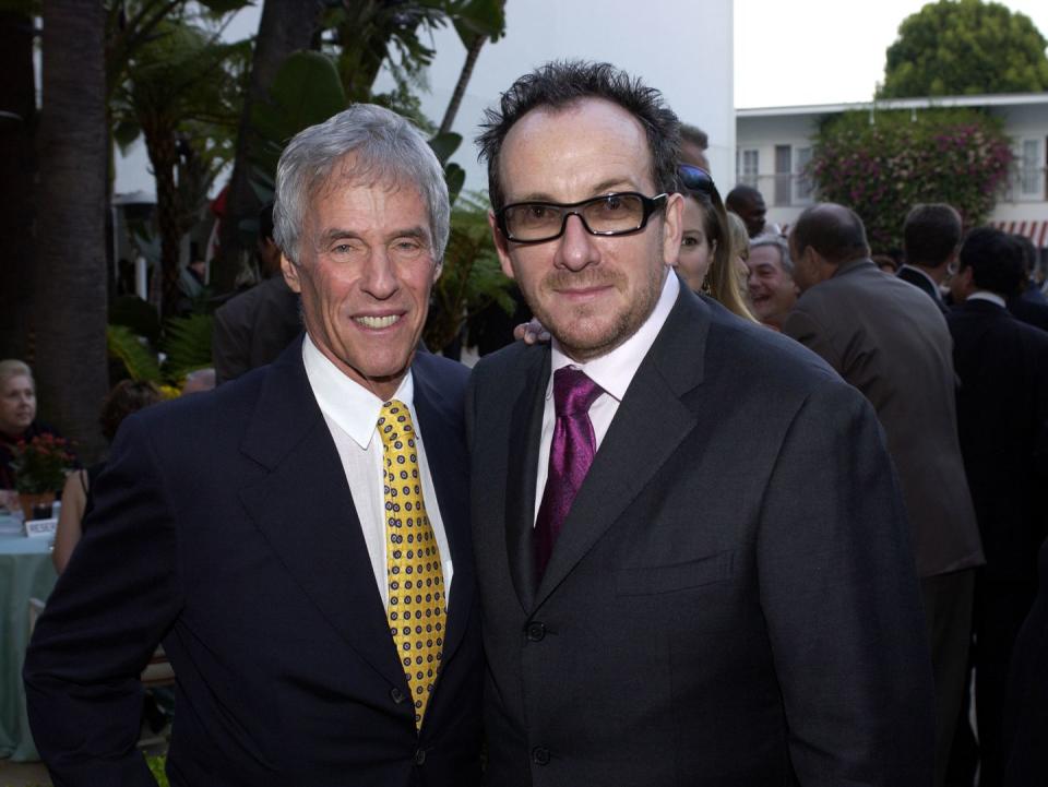 burt bacharach and elvis costello pose for a photo together, each with an around around the other, bacharach is wearing a navy suit coat, white collared shirt, and yellow and blue tie, while costello wears a black suit coat, white collared shirt, a purple tie, and tinted glasses with black frames, both men smile at the camera