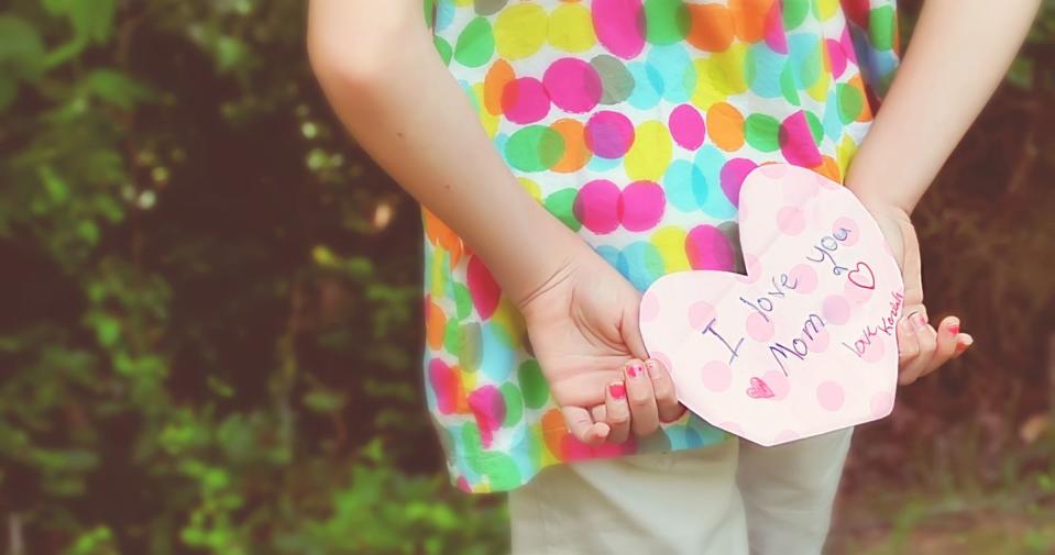 young girl wearing colorful polka dot top holding heart shaped mother’s day card behind her back that says i love you mom