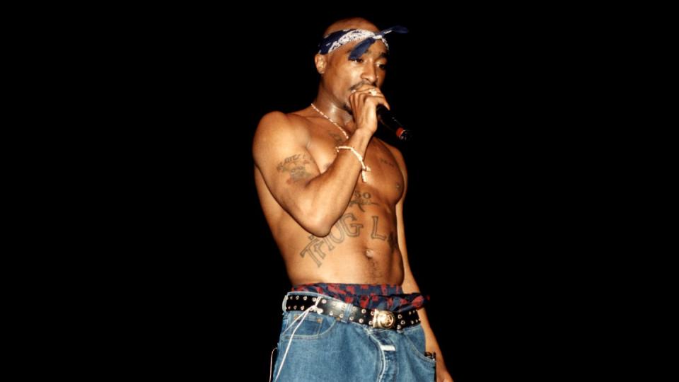 tupac shakur wearing no shirt and jeans, and a bandana on his head, singing into a microphone on a darkened stage