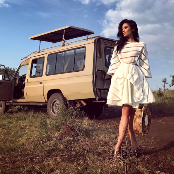 Shay Mitchell in a white dress and heels on location next to a safari vehicle.