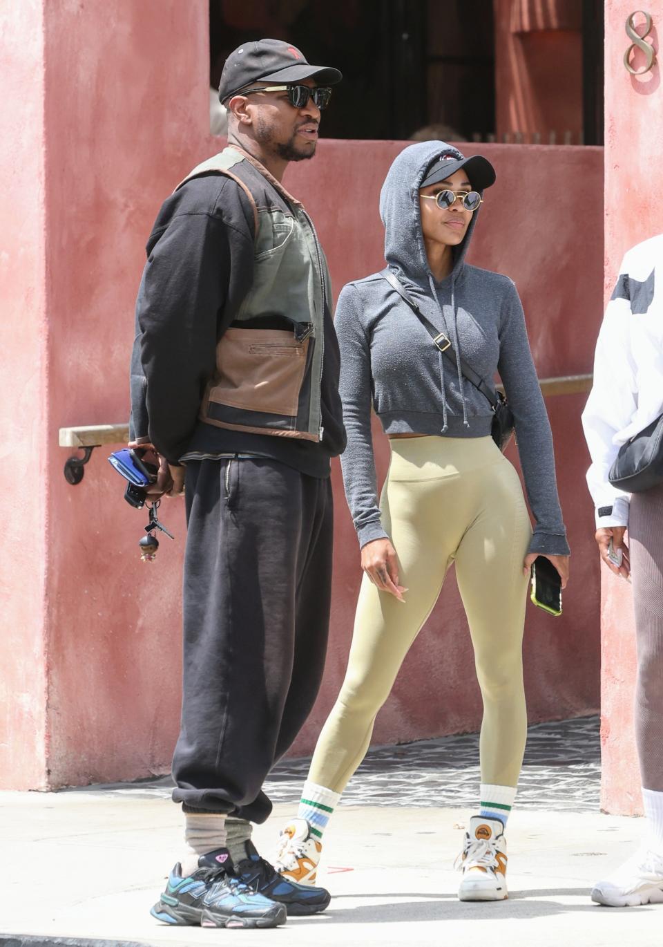 Jonathan Majors and Meagan Good stand outdoors, both in casual wear. Kanye wears a cap, hoodie, and cargo vest. Bianca wears a hoodie, sunglasses, and beige leggings