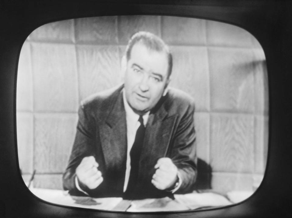 A white man sits behind a desk and is speaking in front of a telivision camera.