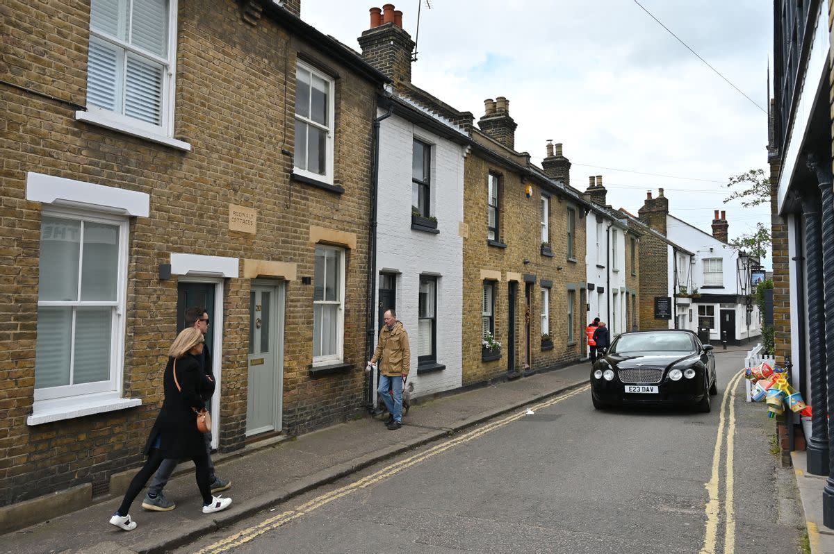 People walk on a street next to a row of homes. A black car drives on the street next to them. 