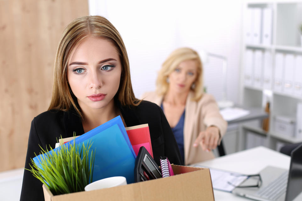 Woman with sad expression carrying cardboard box of office supplies while a seated woman behind her looks on