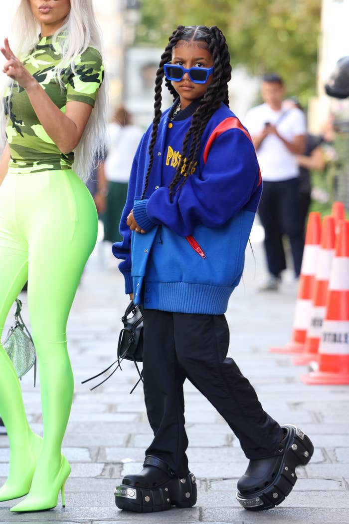 Child in oversized blue sweater, black pants, platform shoes, and sunglasses, standing confidently