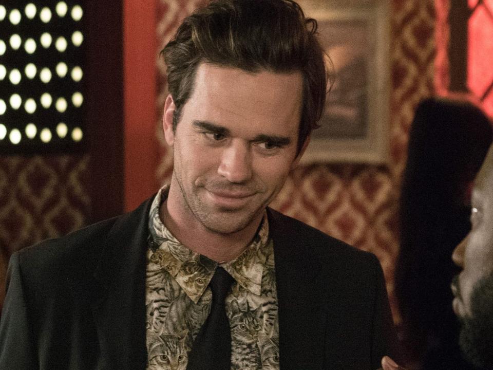 David Walton as Sam wearing a suit with a shirt with brown cats on it holding a cake with cherries