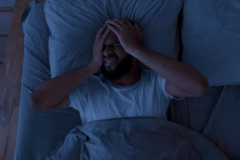 A man is lying in bed, clutching his head with both hands, appearing to be in pain or distress