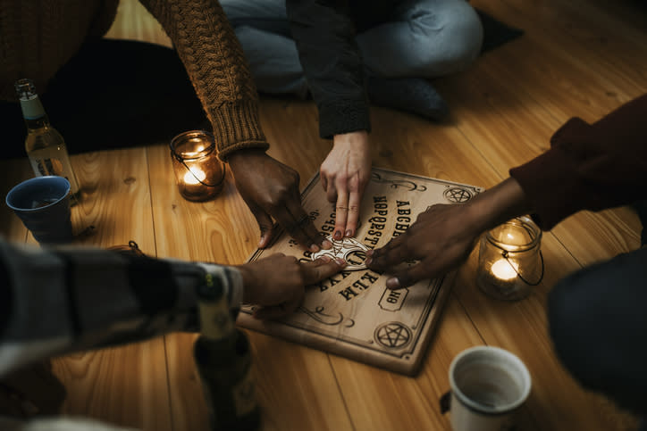 Four people using a Ouija board on a wooden floor, surrounded by candles and drinks