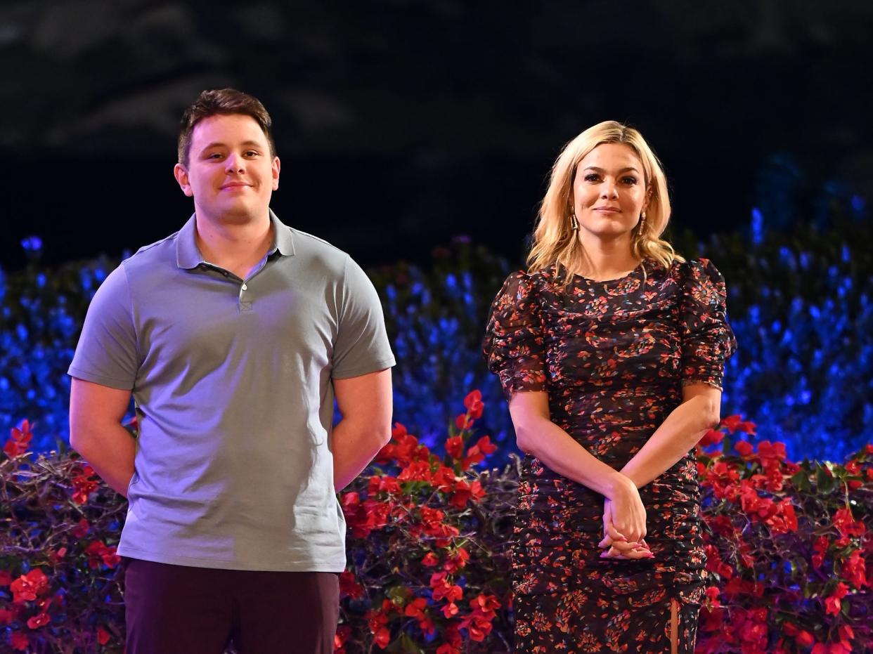 hugo and carly on claim to fame, standing at the top of a small amphitheater next to each other. they're both pensive and look a bit nervous