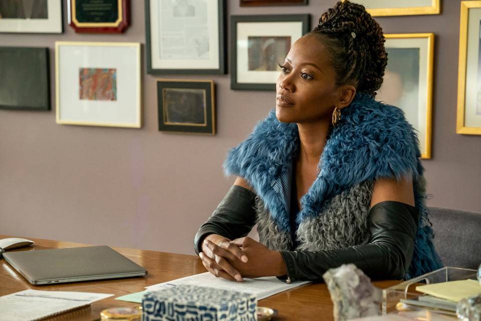 A woman in a blue fur stole sits at a desk in front of a gallery wall.