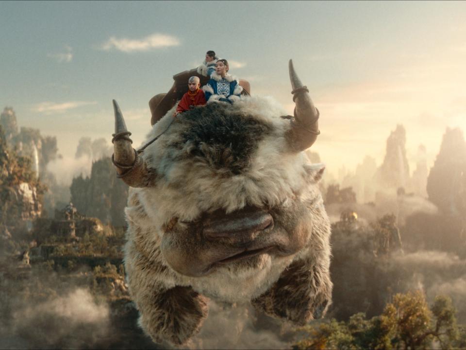 aang, katara, and sokka, teens clad in brightly colored clothing, sitting on top of appa, a large flying bison with fluffy hair, horns, and an arrow pattern on his forehead