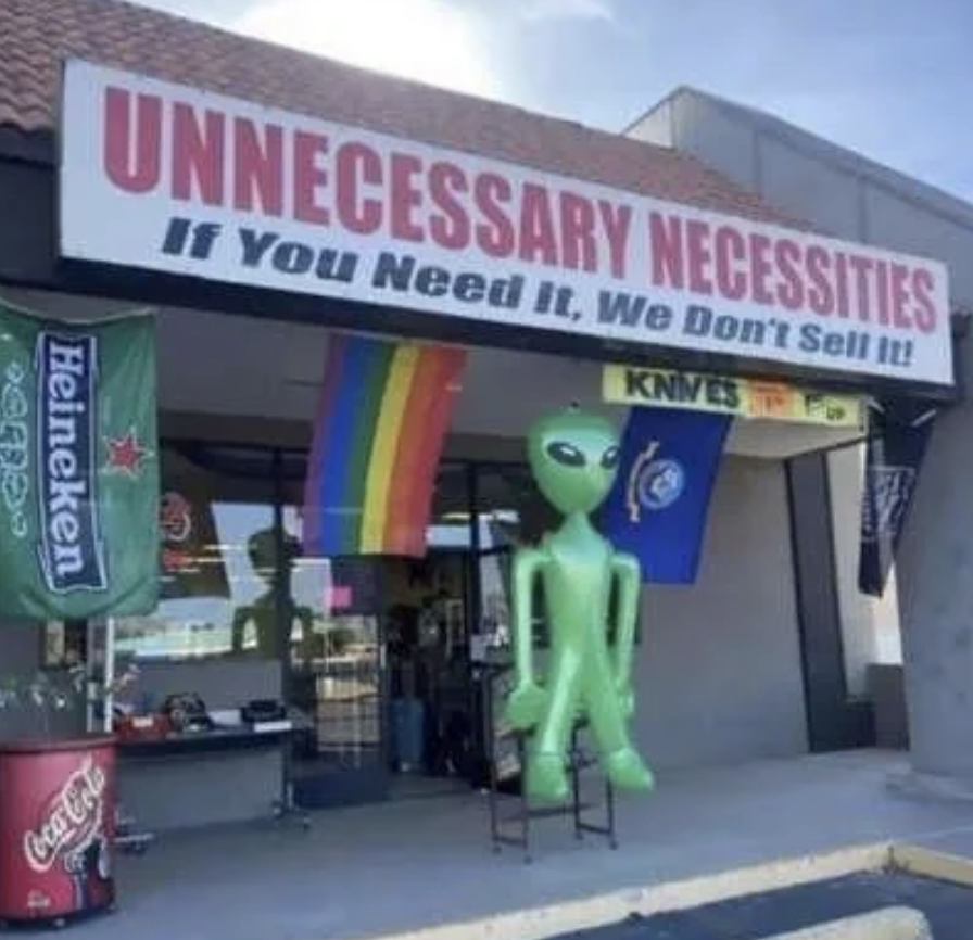 Storefront with a sign reading "UNNECESSARY NECESSITIES: If You Need It, We Don't Sell It!" An inflatable green alien, rainbow flag, and various banners are displayed