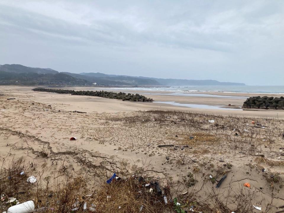 wide beach with dry grass extending to bare sand with a wall of black barriers with more bare sand beyond with puddles of water here and there before reaching the ocean