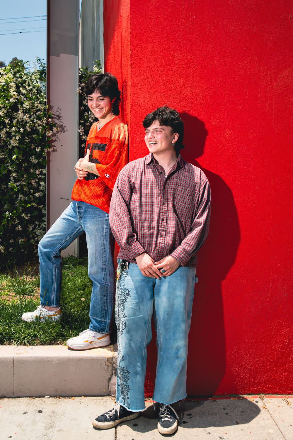 Two of the organizers of Trans Joy Fest stand outdoors next to a red wall..