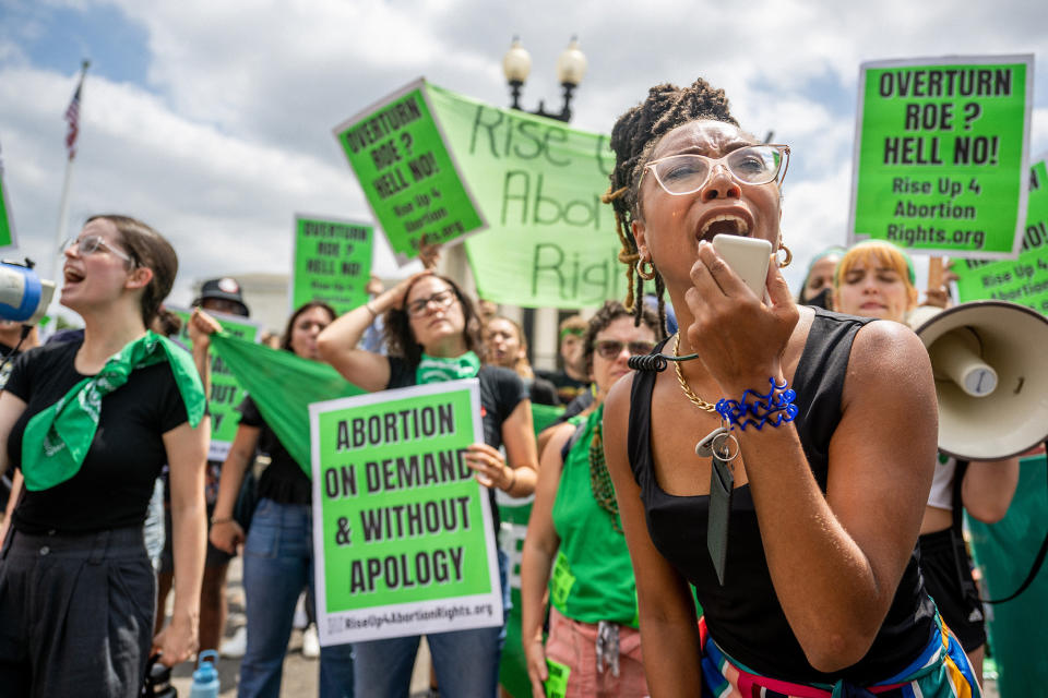 An activist shouts into the microphone of a megaphone, in front of others holding signs reading "abortion on demand and without apology"