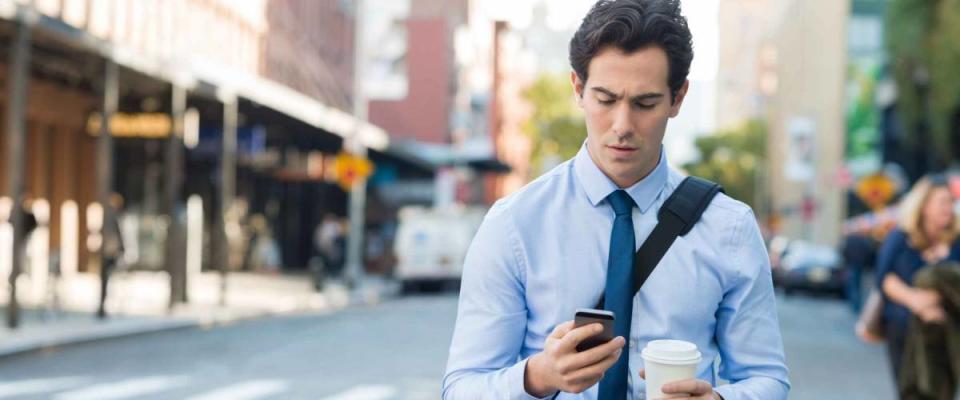 Businessman using smartphone and holding paper cup ina urban scene. Worried businessman walking on the road and messaging with phone.